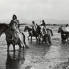 Ute Indians in native dress riding their horses through river.