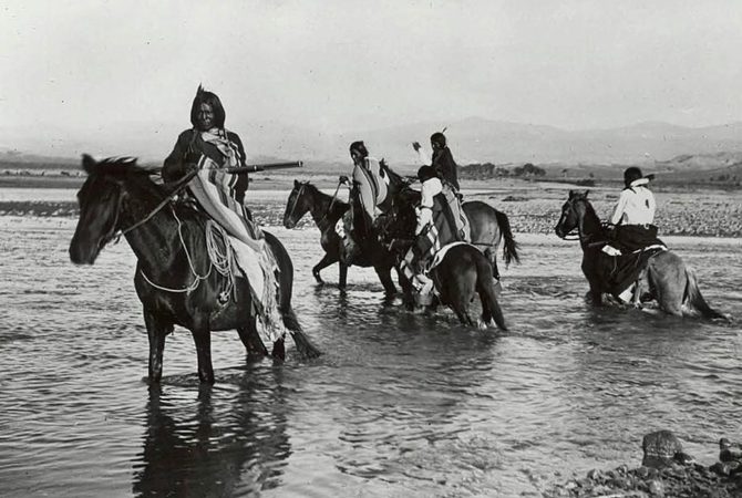 Ute Indians in native dress riding their horses through river.