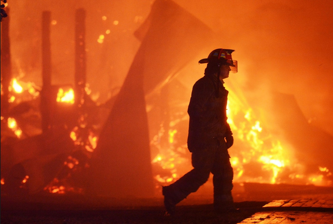 Fire fighter walking by a barn on fire.